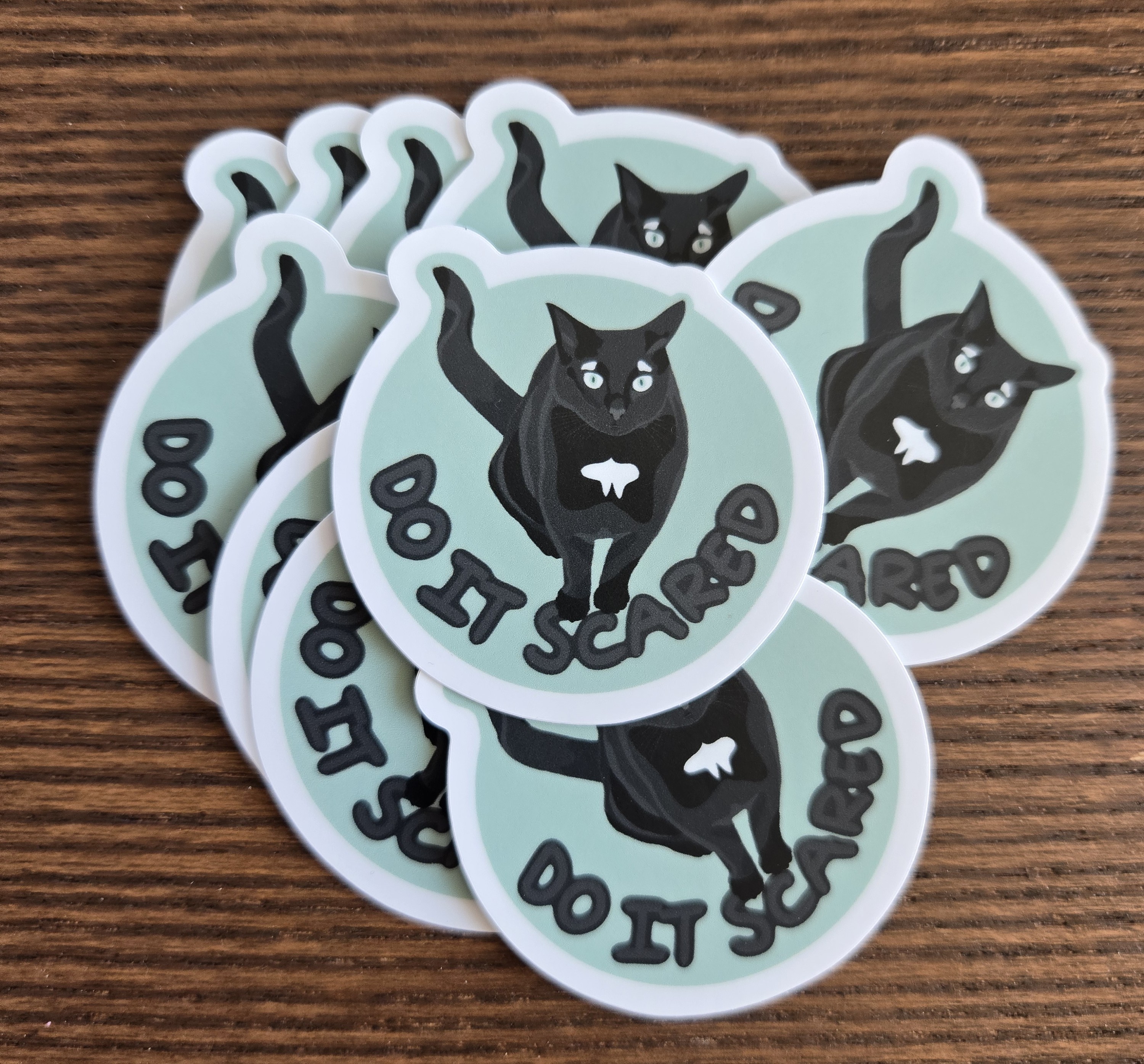 stickers on a table, the sticker is of a black cat with airplane ears with the text DO IT SCARED underneath