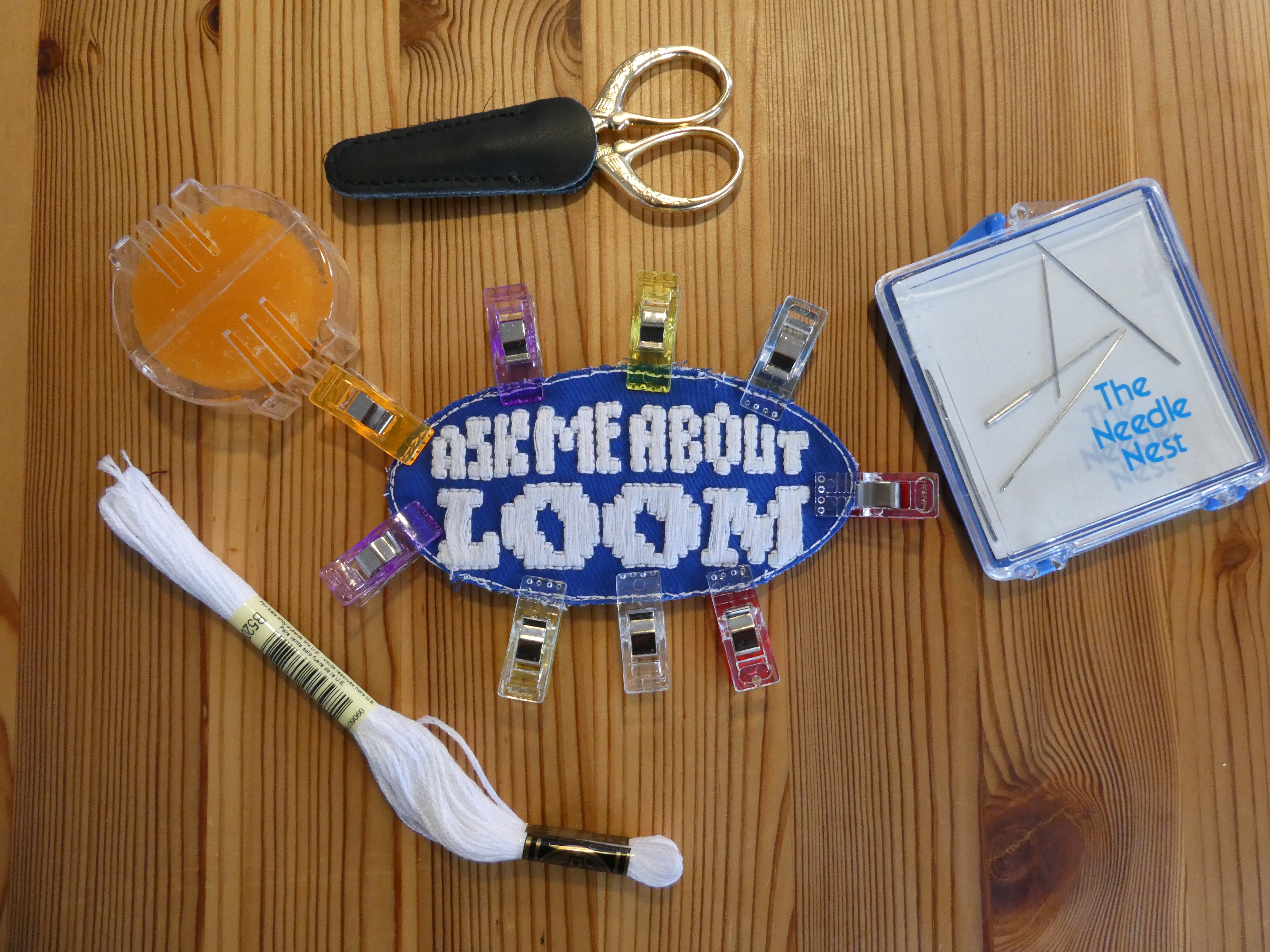 a patch on a blue background that says 'ask me about LOOM' with various embroidery supplies around it